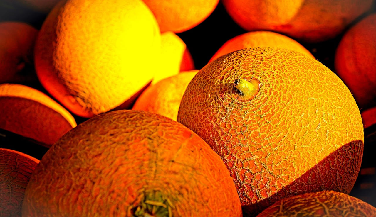 Meloni interi e affettati con polpa arancione esposti al mercato, con foglie verdi attaccate e gocce d'acqua sulla buccia reticolata